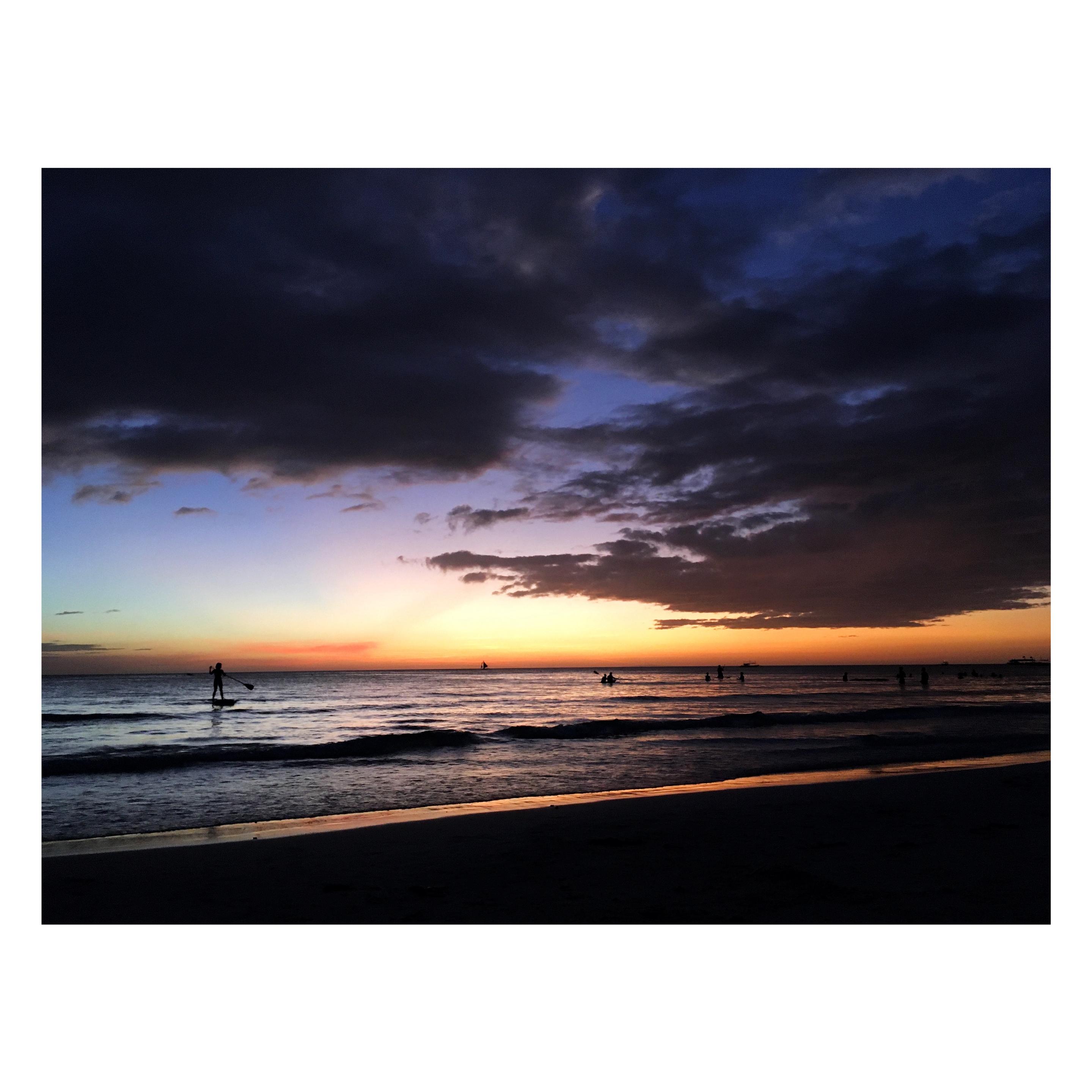 A dramatic sunset on an island beach. Masses of low clouds roll across a deep blue evening sky that transitions to orange and gold where the sun sinks into a tranquil sea. Distant silhouetted figures dot the water, including a person standing on a paddleboard at left and a sailboat on the horizon. The bright diagonal line of the surf glows in the fading light, contrasting with the dark sand in the foreground.

Today’s FotoVorschlag challenge asks us to share our “most beautiful moments” with @undarzt.bsky.social, who is very ill according to his own post. Sir, I may not know you but I send my sympathy and regards. I chose this photo, taken from a trip to the Philippines in 2020, because it made me think of a fact I had been reminded of recently: that the light that touches us captures our likeness and travels onward, forever. Just as starlight that travels a million light-years to reach our eyes allows us to see a star as it was a million years ago, so too does the light reflecting off