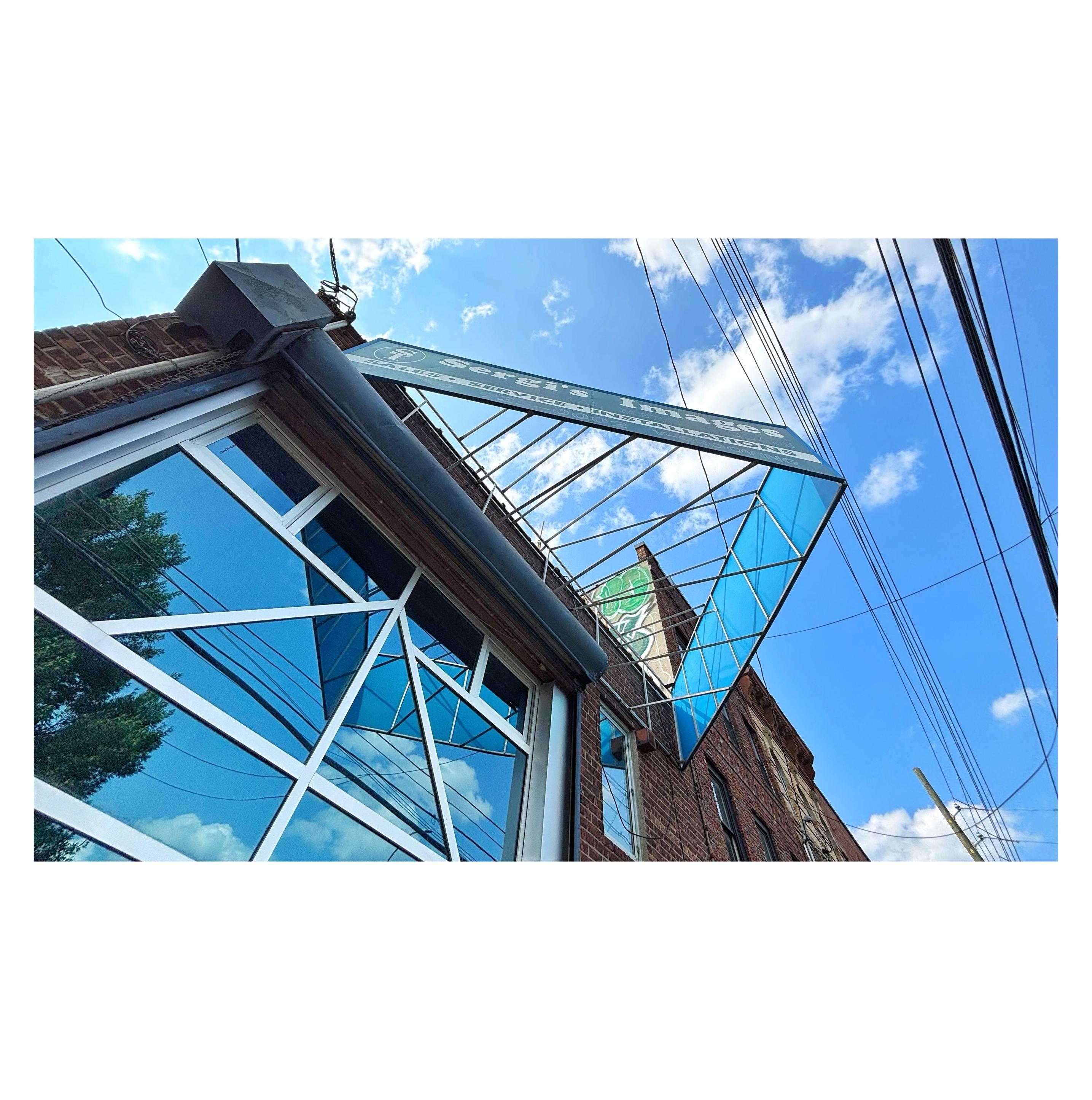Angled view looking up at a brick building facade under a blue sky with white clouds. A storefront sign projects outward from the building, its two ends attached to the facade while its midpoint extends out furthest, so that it can be more easily read from the sidewalk and creates a triangle through which we can see the sky. The visible side reads “Sergi’s Images” and I presume the other side says the same thing. Underneath is a large window made of triangular and trapezoidal panels that reflect the sky and a tree across the street, as well as, most notably, the triangle made by the sign — in effect creating a square out of the sign’s actual and reflected sides. Power cables crisscross the sky overhead. The image is striking for its geometric lines and shapes.