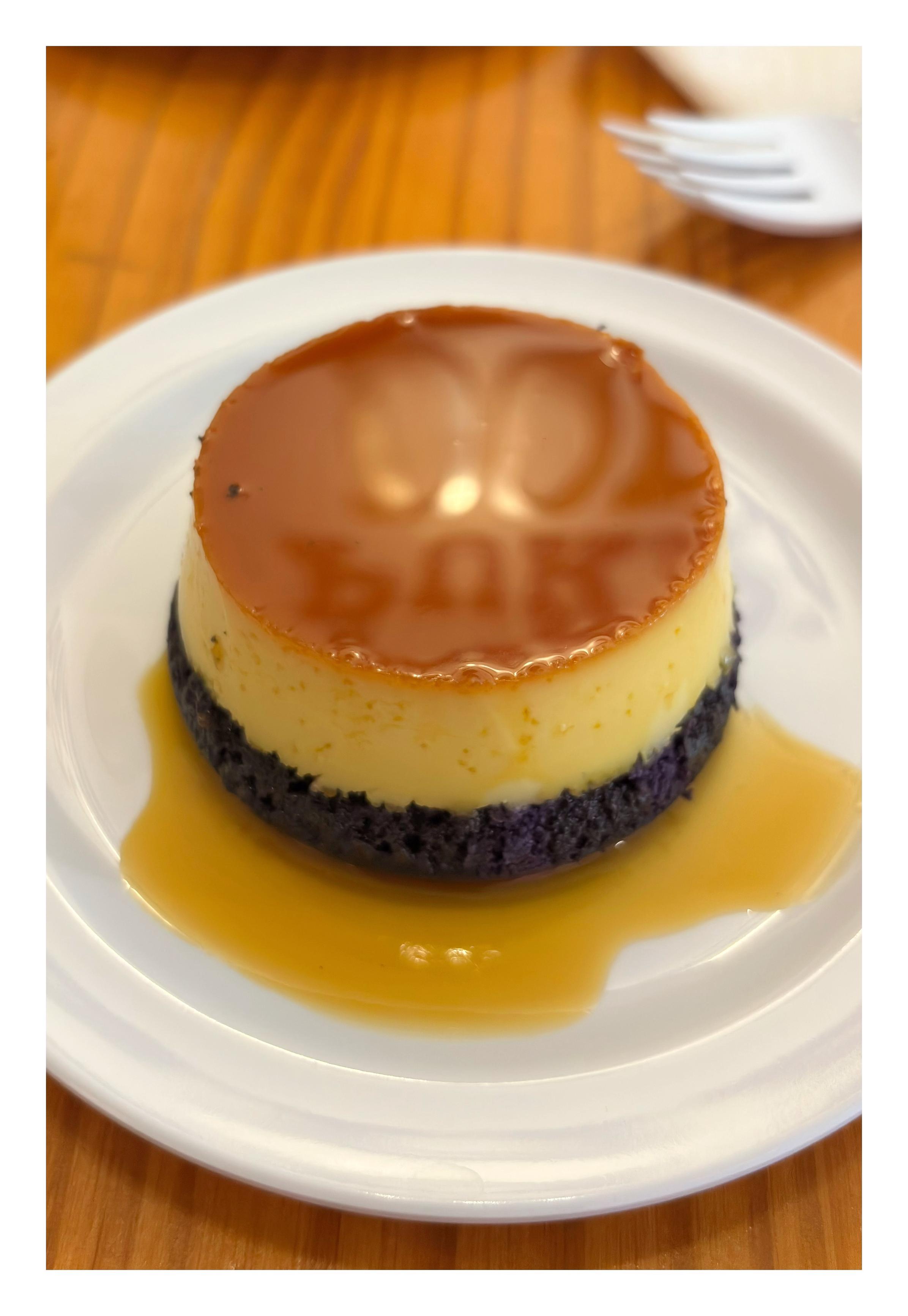 Closeup of a circular serving of leche flan on a white plate, atop a wooden table with some blurred plastic forks in the background. Leche flan is a traditional Filipino sweet custard with a glazed topping of liquid caramel, in this case with a thin base of cake flavored with ube (purple yam). The custard sits in a pool of syrup, and the caramel glaze on top partially reflects the “Purple Dough” logo mounted on the opposite wall.