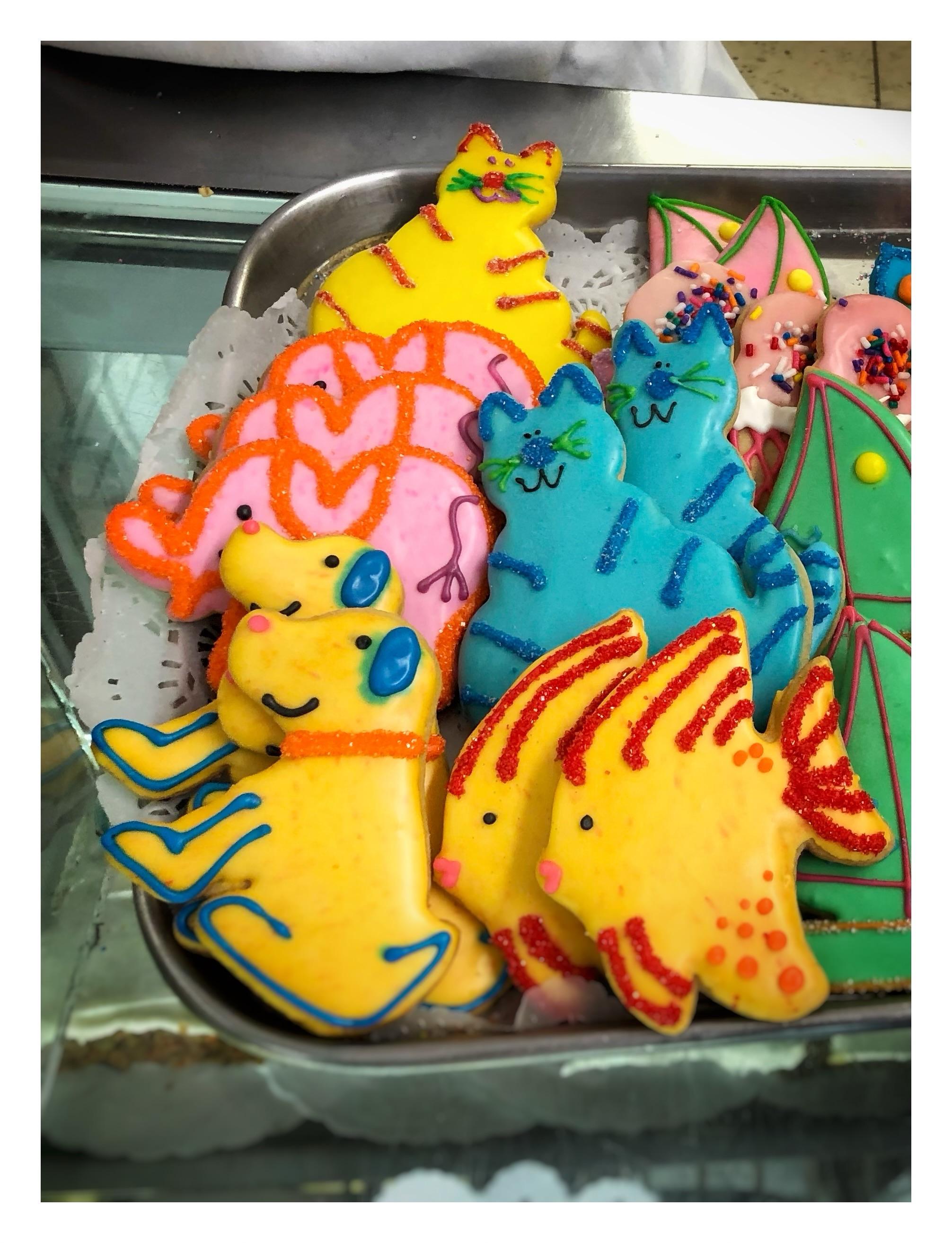 Closeup of a metal tray of cookies cut into animal shapes and decorated with multicolored glaze and icing: smiling cats, dogs, elephants, and angelfish, among others.