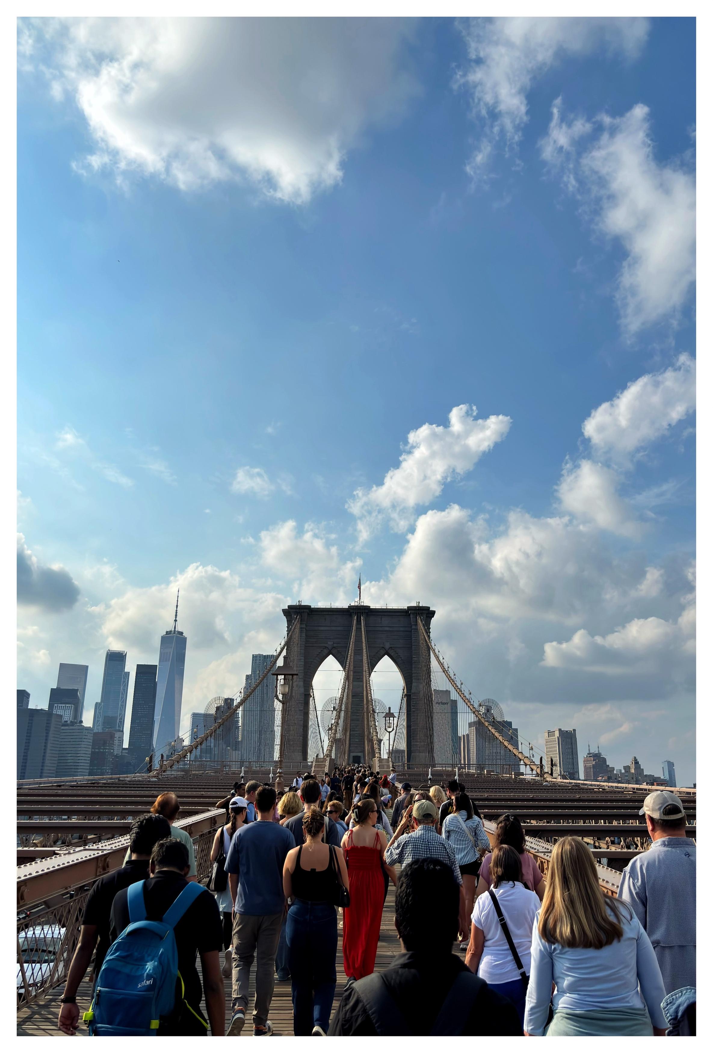 A view of the eastern tower of the Brooklyn Bridge as seen from its pedestrian walkway, facing the Manhattan side. Behind it is the Manhattan skyline under a wide blue sky with cottony clouds. A throng of people facing away from camera walks across the bridge, most notably a woman in the middle of the crowd whose vivid red spaghetti-strap dress catches the eye.