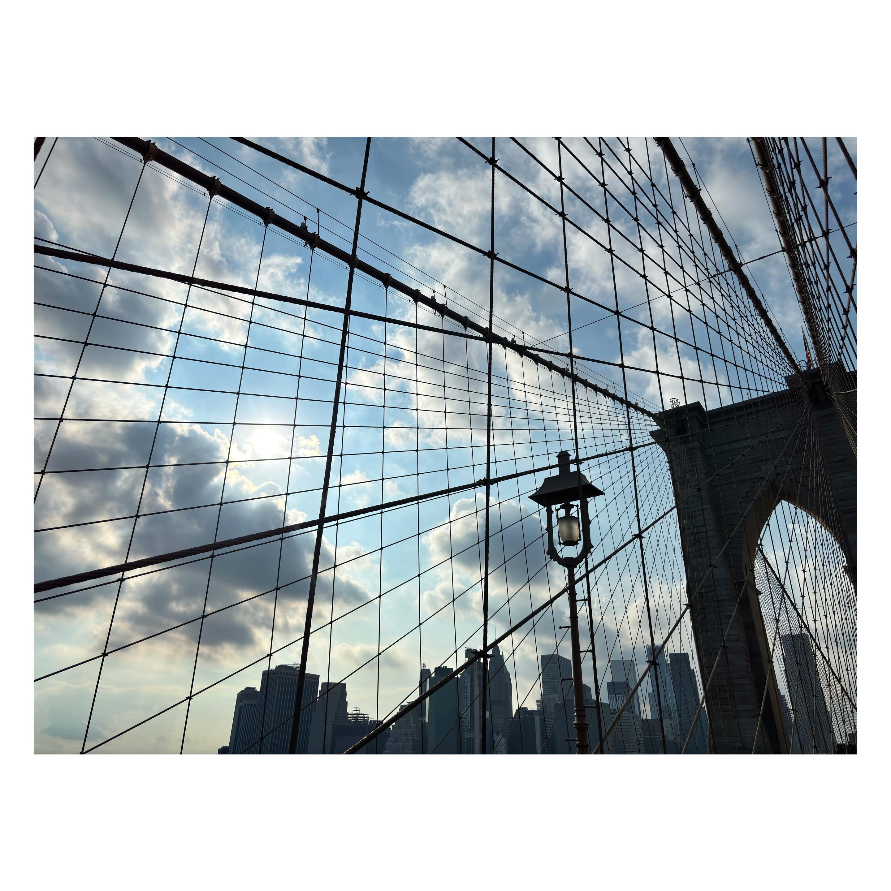 A view of the Manhattan skyline under a blue sky, with the sun shining through white clouds, as seen through the network of crisscrossing cables radiating out from one of the towers of the Brooklyn Bridge at right. In the foreground is an old-fashioned lantern on a post standing alongside the bridge’s pedestrian path.