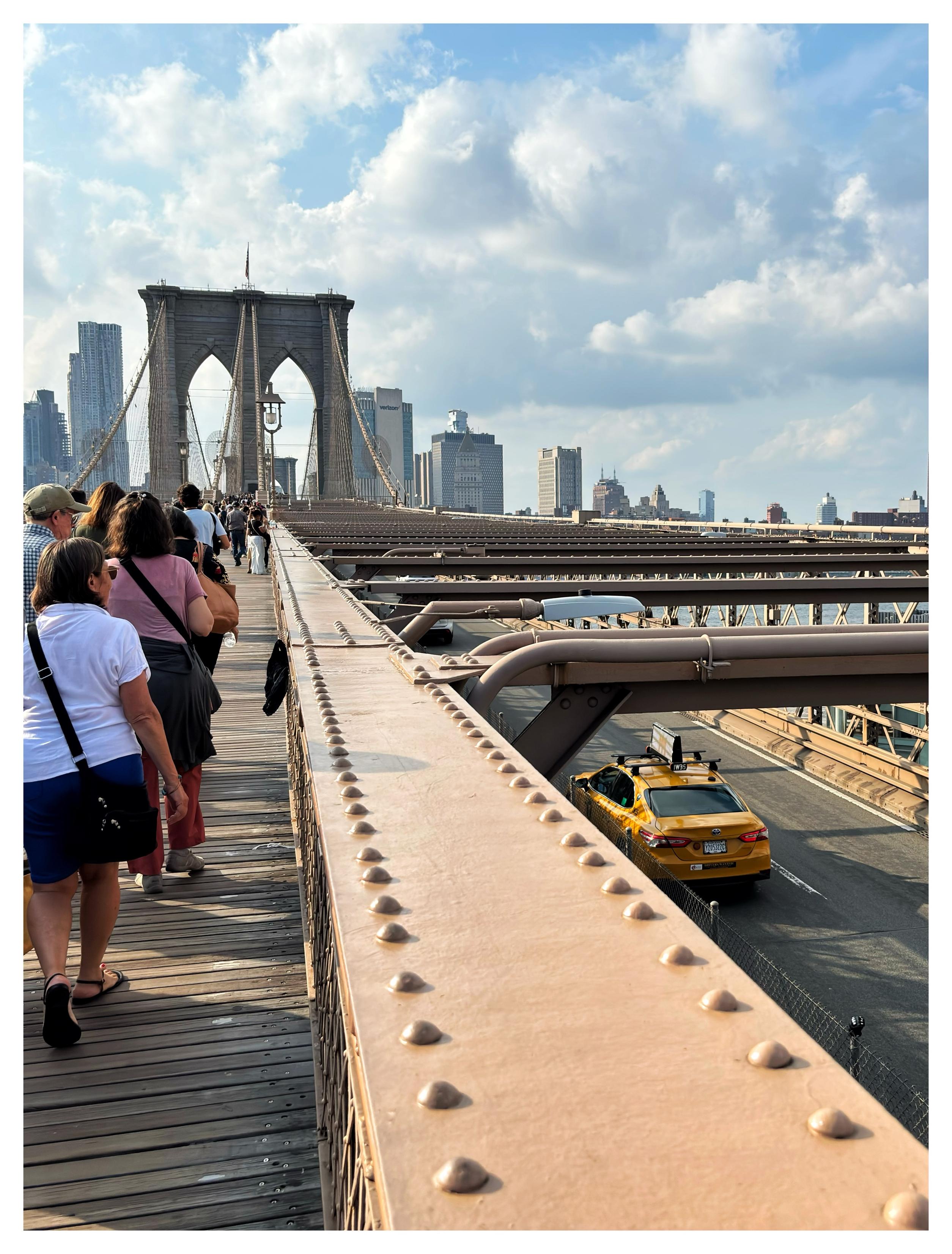 A view from the pedestrian walkway of the Brooklyn Bridge, facing the Manhattan skyline under a blue sky with masses of white cumulus clouds. People facing away from camera walk along the path of wooden planks at left. To their right is a wide raised metal divider studded with two rows of rivets, all receding away to the double-arched bridge tower in the distance. Below and to the right of the divider is the bridge’s roadway, where we can see a yellow cab speeding off towards Manhattan.