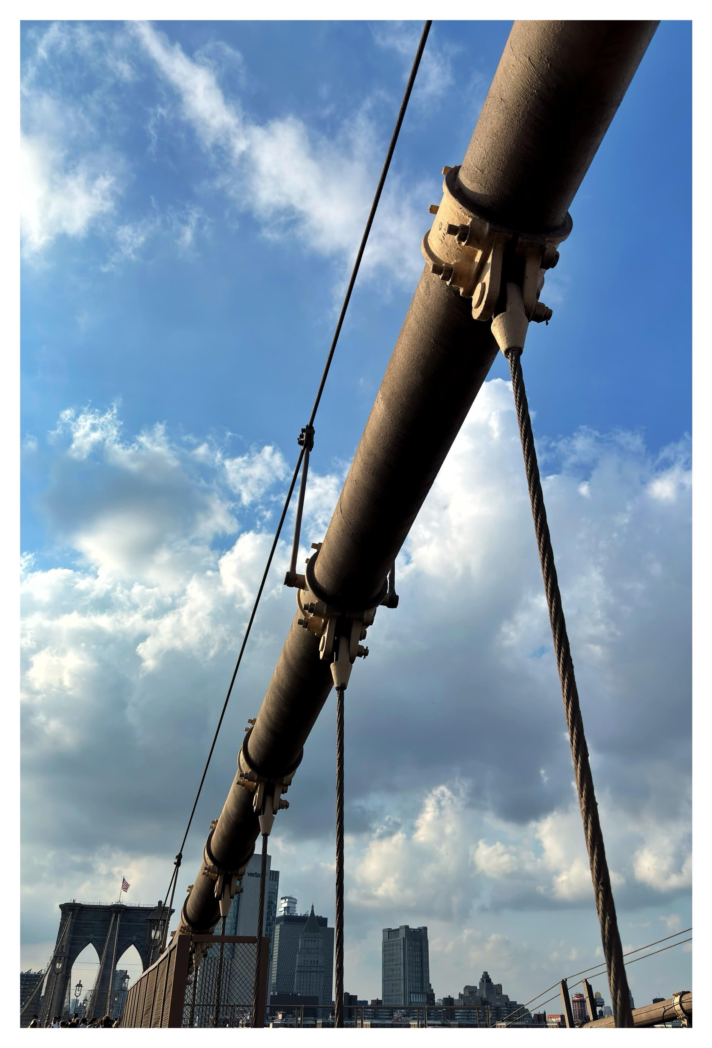 A view from underneath one of the massive main cables of the Brooklyn Bridge, soaring diagonally across a blue sky with white clouds towards one of the double-arched bridge towers in the distance. Suspender ropes hang vertically from the main cable, holding up the bridge’s trusses out of frame below.
