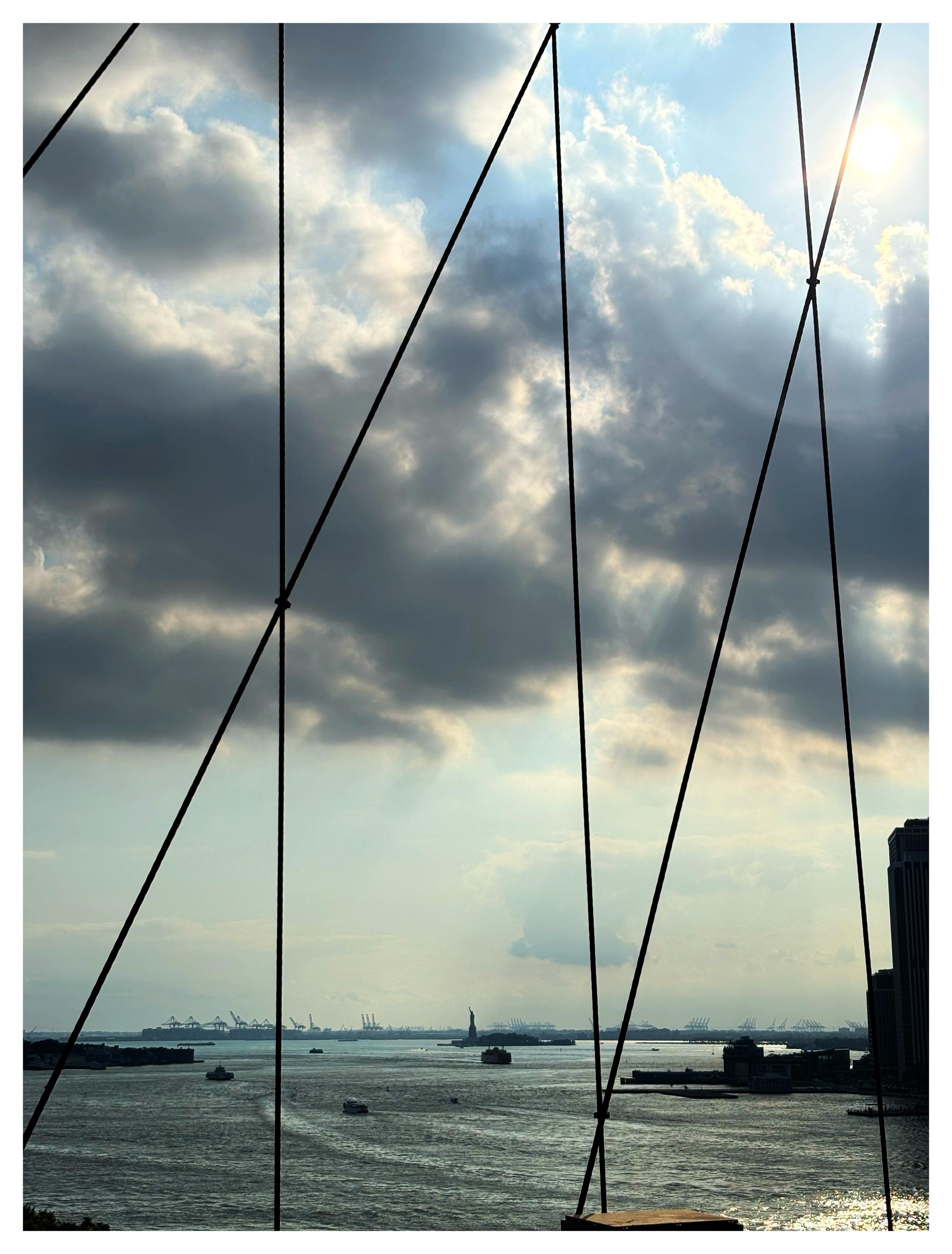 New York Harbor as seen from the pedestrian walkway of the Brooklyn Bridge, with the bridge’s thin crisscrossing cables chopping up the view into trapezoidal sections. Through the cables we see the wide waters of the bay, dotted with boats, under a heavily clouded sky with the sun shining through at upper right. In the far distance is the tiny Statue of Liberty, and beyond it, a line of shipping cranes on the New Jersey shore at the horizon.
