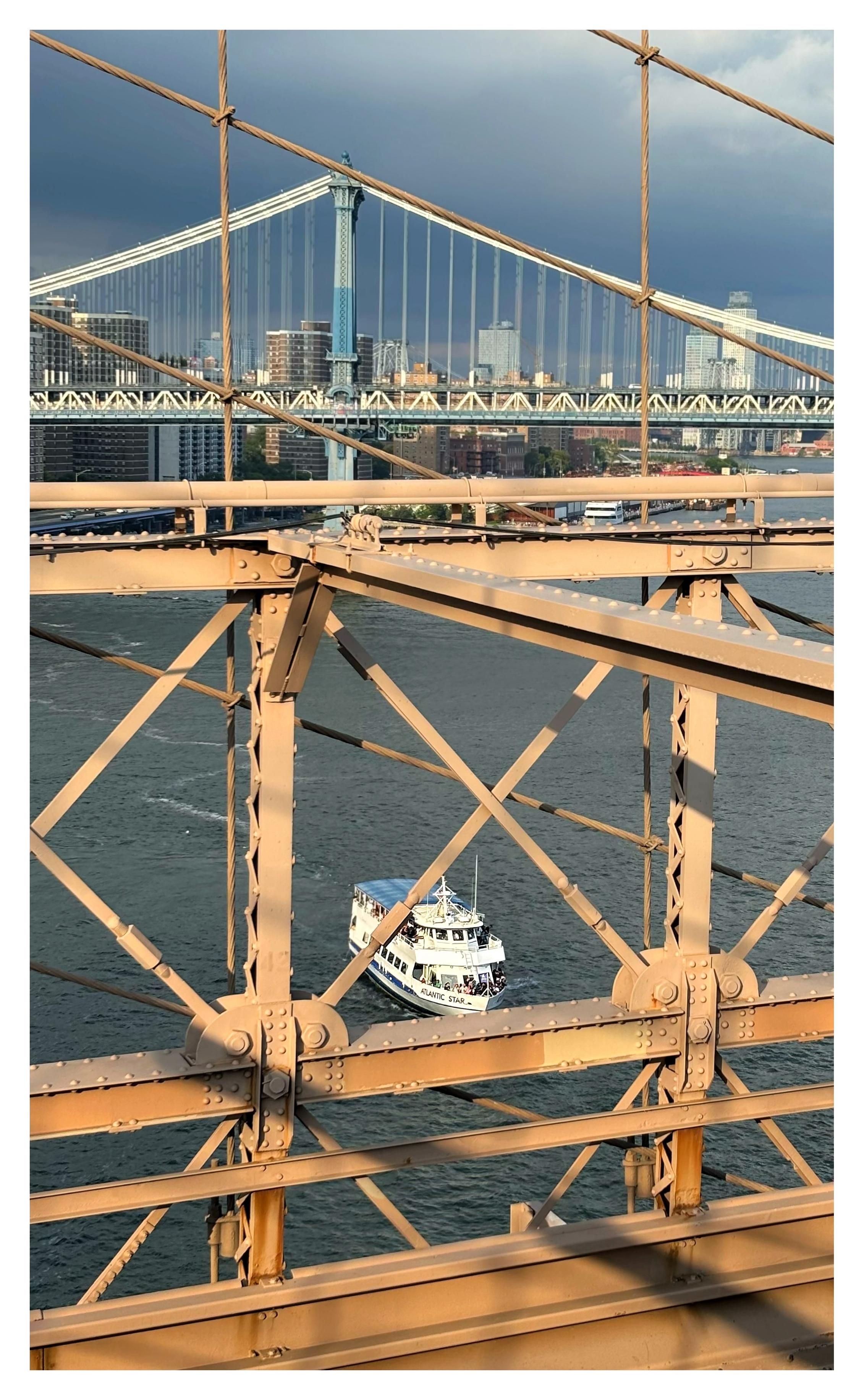 A view of the East River from the pedestrian walkway of the Brooklyn Bridge, looking down through the metal beams of its truss. In between the beams we see a white double-decked ferry, the Atlantic Star, moving through the water as it prepares to cross under the bridge. Above and through the bridge’s suspension cables we can see the Manhattan Bridge in the distance, against a backdrop of tall buildings under an overcast sky.