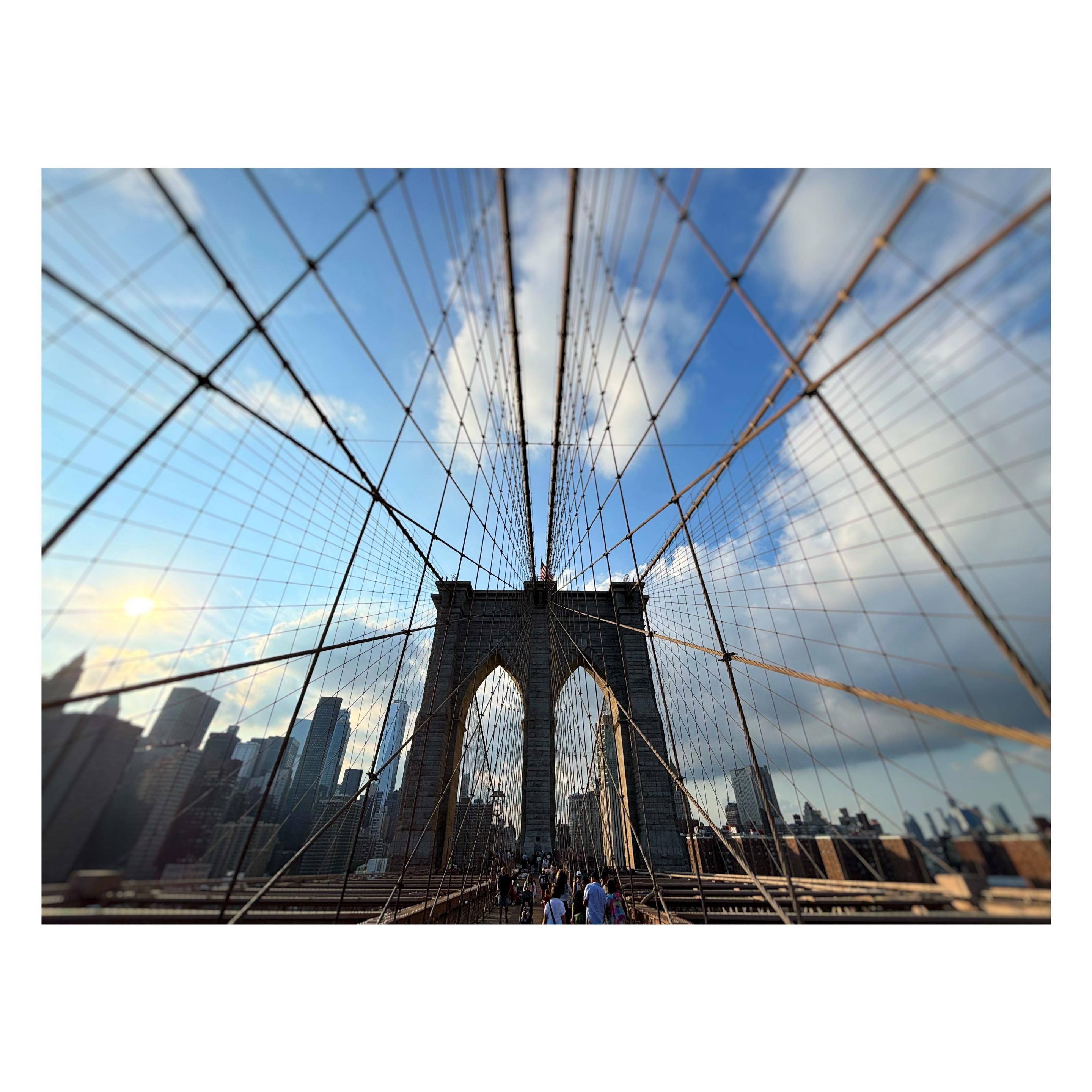 Wide-angle view of one of the double-arched towers of the Brooklyn Bridge. A crowd of people walks along the pedestrian pathway below. A network of crisscrossing cables radiates from the tower like a spiderweb, shooting out to the extreme foreground at left and right. Beyond the bridge tower is the Manhattan skyline under a blue sky with clouds. The image is sharply in focus at the tower but blurred around the edges.