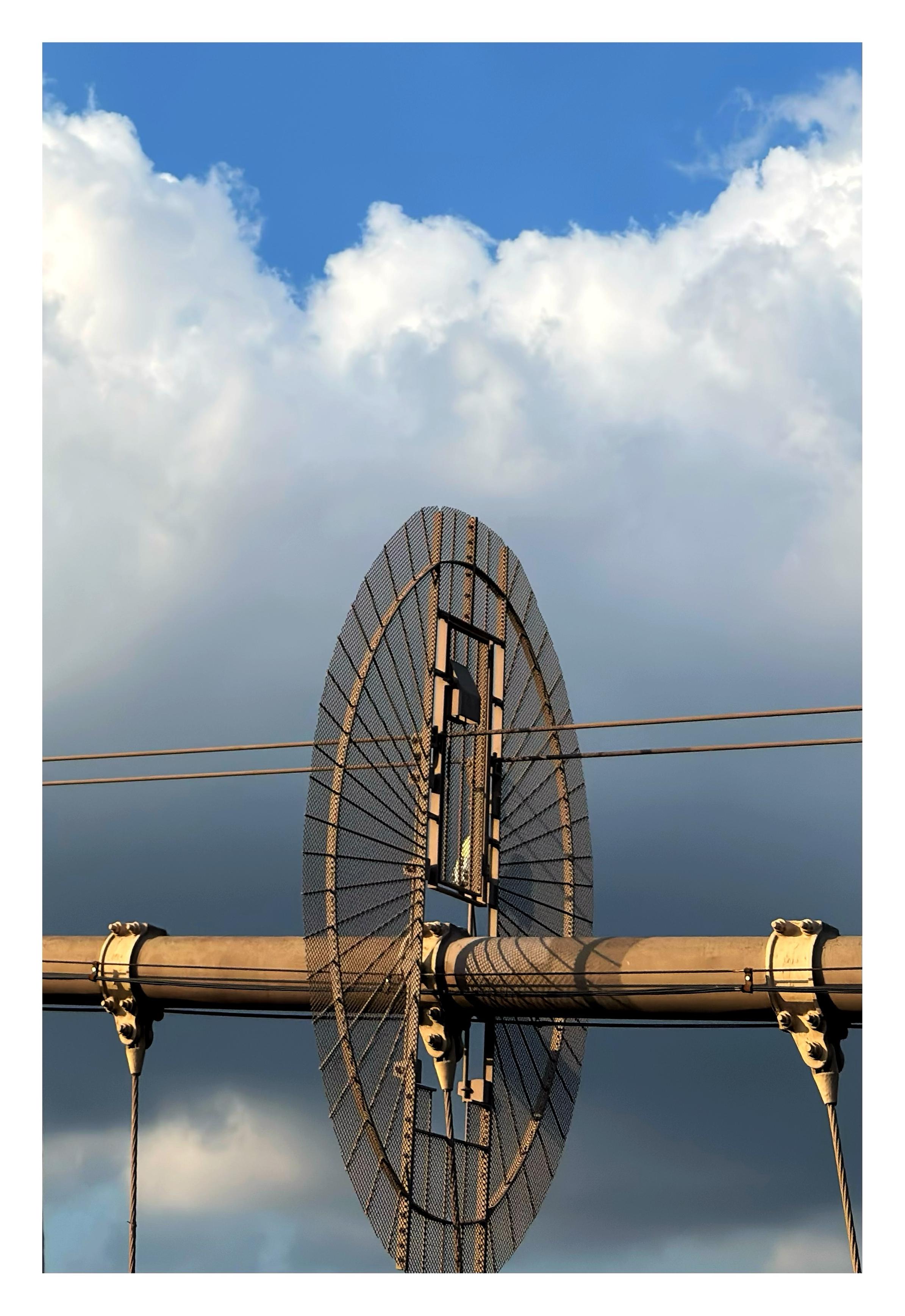 A near-horizontal section of one of the main suspension cables of the Brooklyn Bridge, against a backdrop of massive white clouds with a bit of blue sky at the top. Around the middle of the cable is a large circular metal screen barrier, oriented vertically, with spokes radiating out from its center. A closed screen door is set into its upper half.