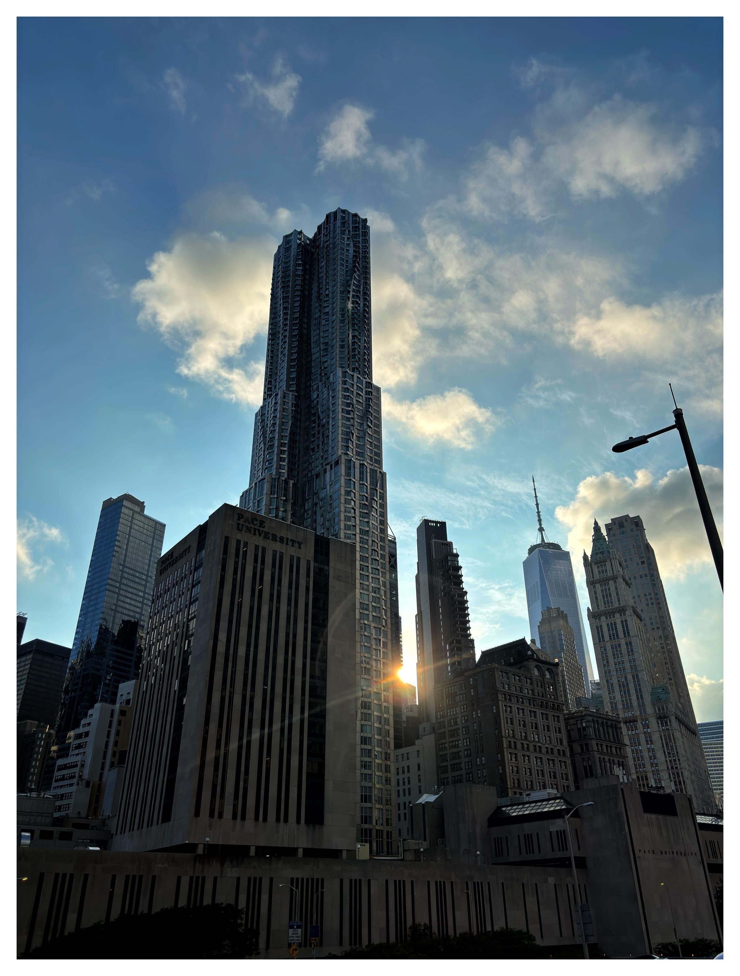 Two photos featuring the sun in different settings, with different backlit elements in the foreground. In this first photo, the afternoon sun shines in the narrow space between two tall buildings in a Manhattan cityscape, under a blue sky with white clouds. I shot this as I was walking across the Brooklyn Bridge.