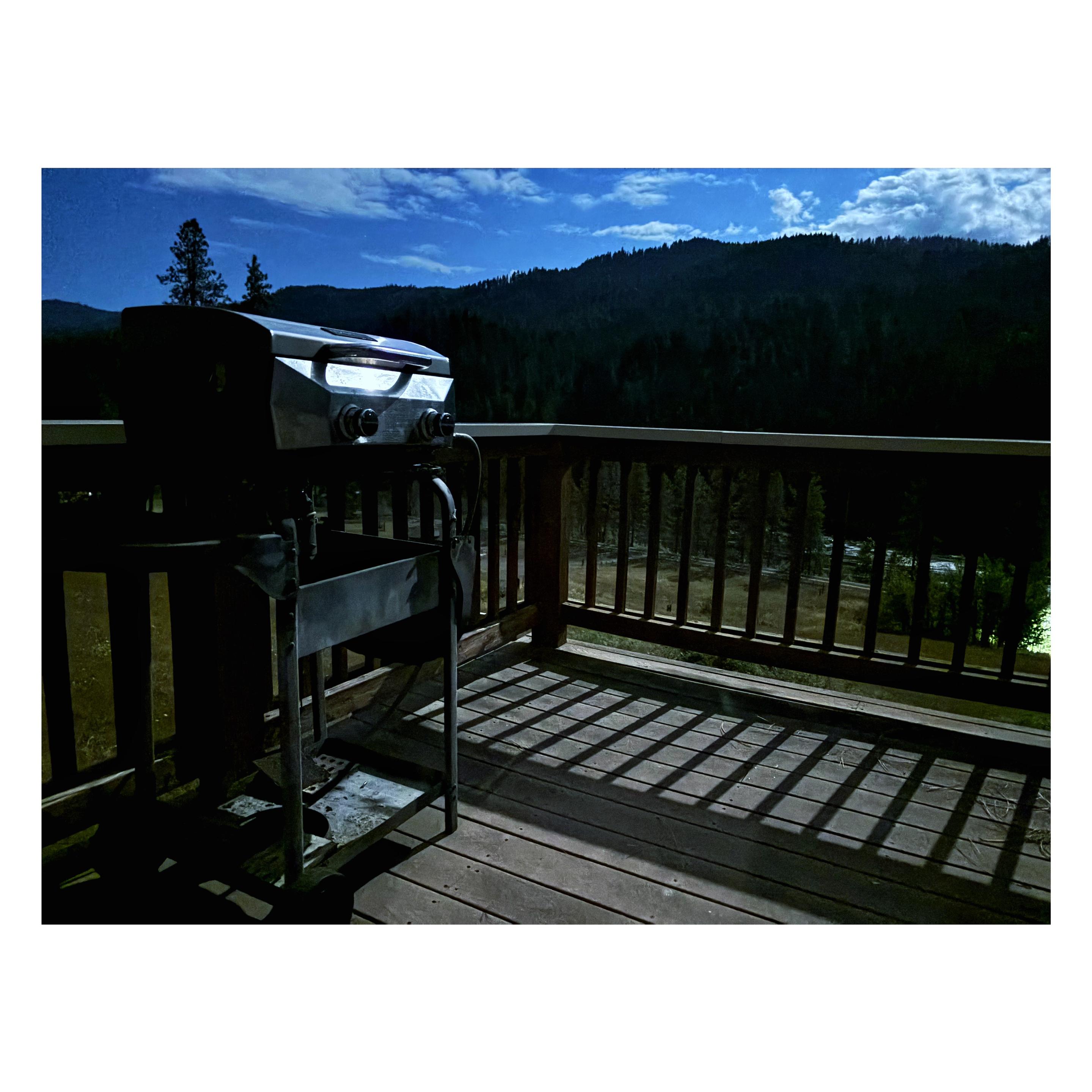 An old barbecue grill sits at the side of a wooden deck with a guardrail of vertical posts. Beyond is a field sloping down to a dark conifer forest under a deep blue evening sky with clouds. The scene is illuminated solely by moonlight, which gleams off the grill and throws the guardrail’s striped shadows onto the deck.