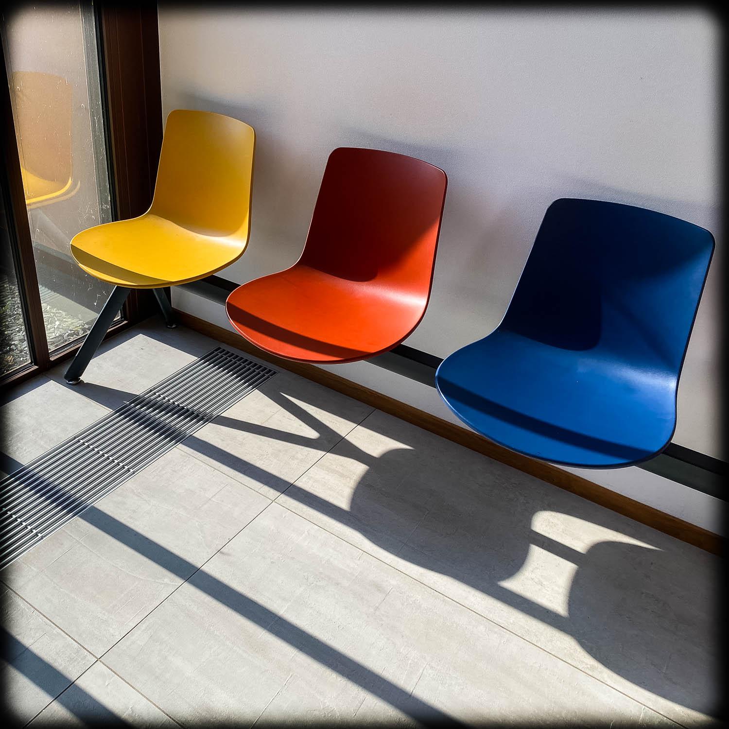 Photograph of a row of three plastic chairs, yellow, red and blue, lit from the side with strong shadows from a window.