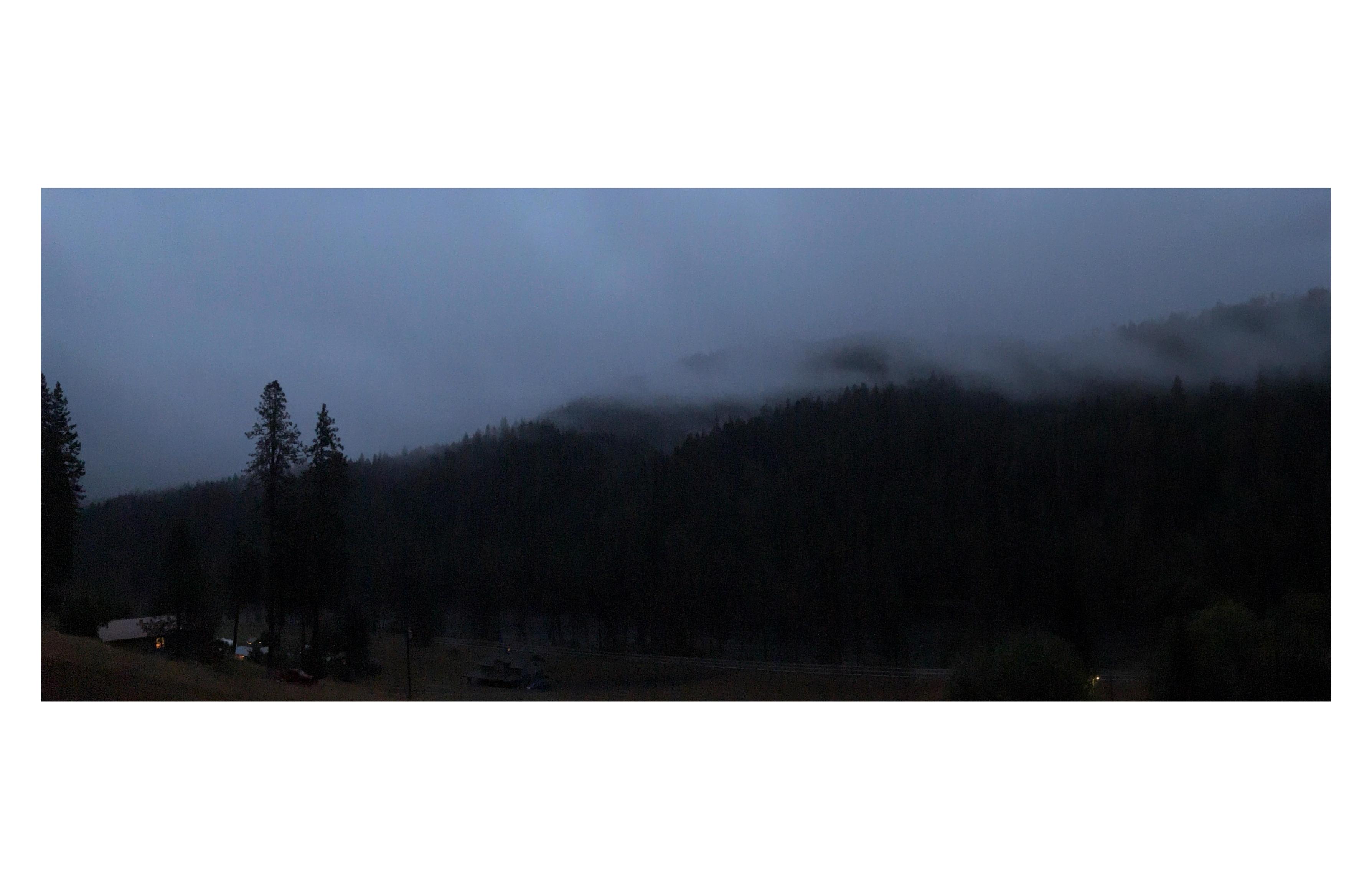 Third in a series, taken in the evening the next day. The landscape is dark and indistinct, the forest a wall of shadow, with a low pale fog covering the sky and outlining the treetops. The lit windows of the distant houses are tiny points of light in the field below.