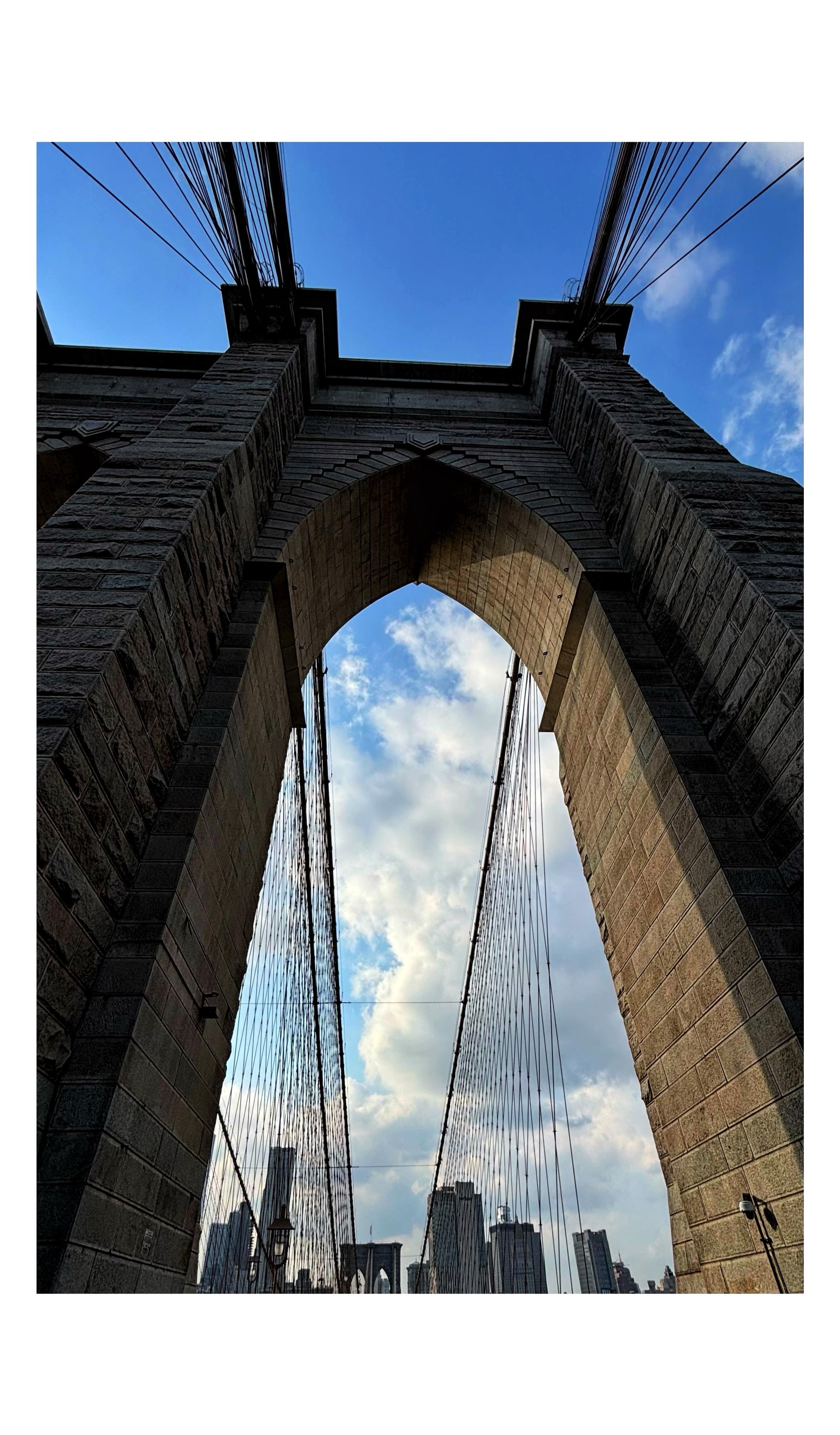 A photo I took looking up at one of the massive limestone-and-granite towers of the Brooklyn Bridge as I was walking on the pedestrian path directly underneath. Through the tall, sunlit archway we see cables stretching out to the second bridge tower in the distance, against the backdrop of the Manhattan skyline under a blue sky with cumulus clouds.
