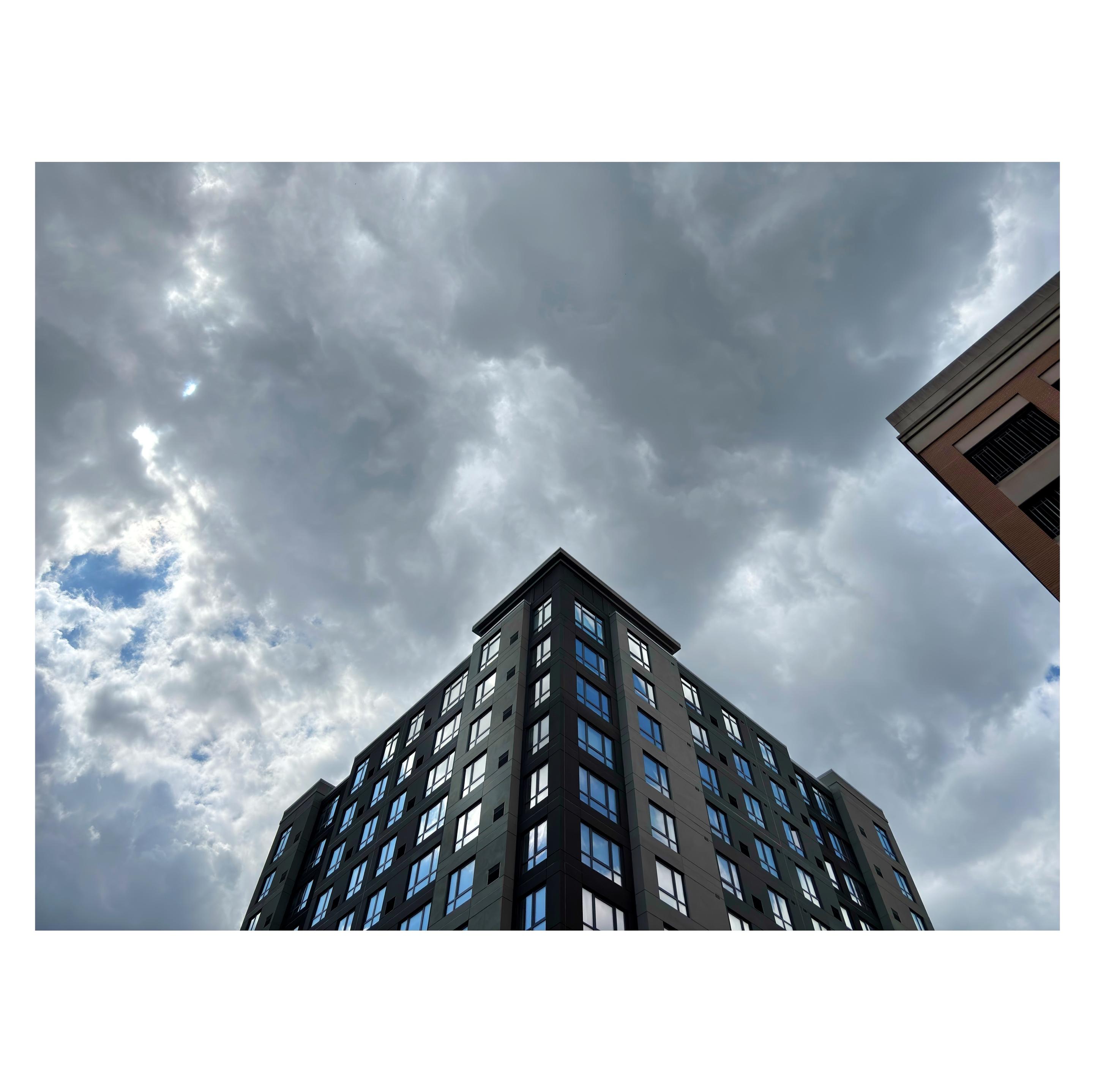 Photo looking up from street level towards the top floors of a high-rise building under an overcast sky. The two visible sides of the building meet at a corner in the middle of the image, giving its roofline a triangular shape. At left, a break in the gray clouds offers a glimpse of blue sky, while the building’s grids of windows reflect the blue sky and clouds behind camera. At right, the top corner of another building pushes in, creating another triangle whose edges are parallel to those of the main building.