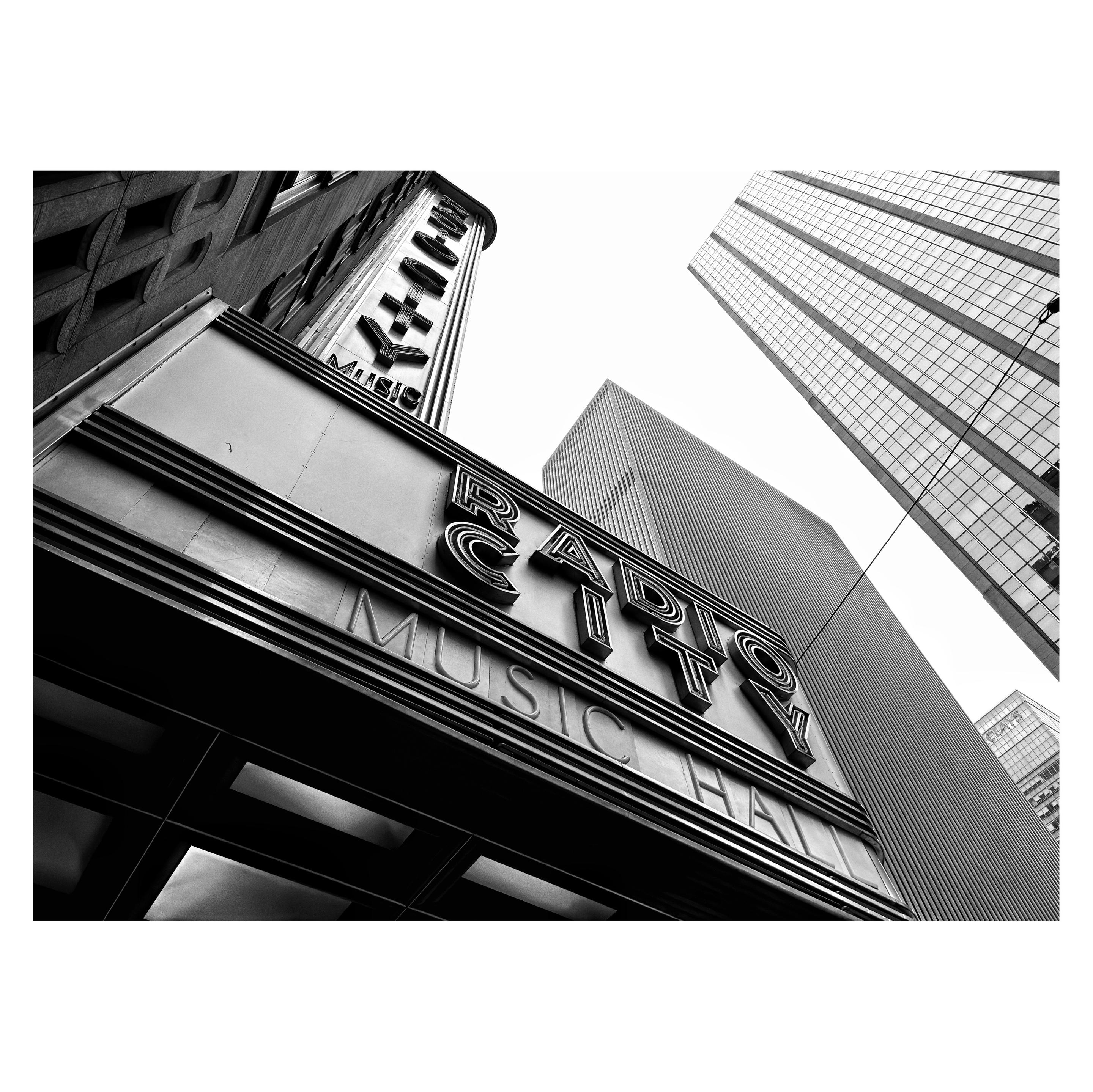Black and white photo looking up at the marquee sign of Radio City Music Hall in Manhattan. The perspective is from almost directly underneath the sign so that the lines and angles of the scene all recede from the viewer at an extreme angle. Closest to camera is the large horizontal marquee that reads “RADIO CITY MUSIC HALL” extending over the sidewalk from the building facade, with some rectangular lighting panels visible underneath. Above it, a vertically oriented sign with the same words is mounted on the side of the building. In the background are the surrounding skyscrapers, all steel and glass and window grids, jutting into a blank white sky.