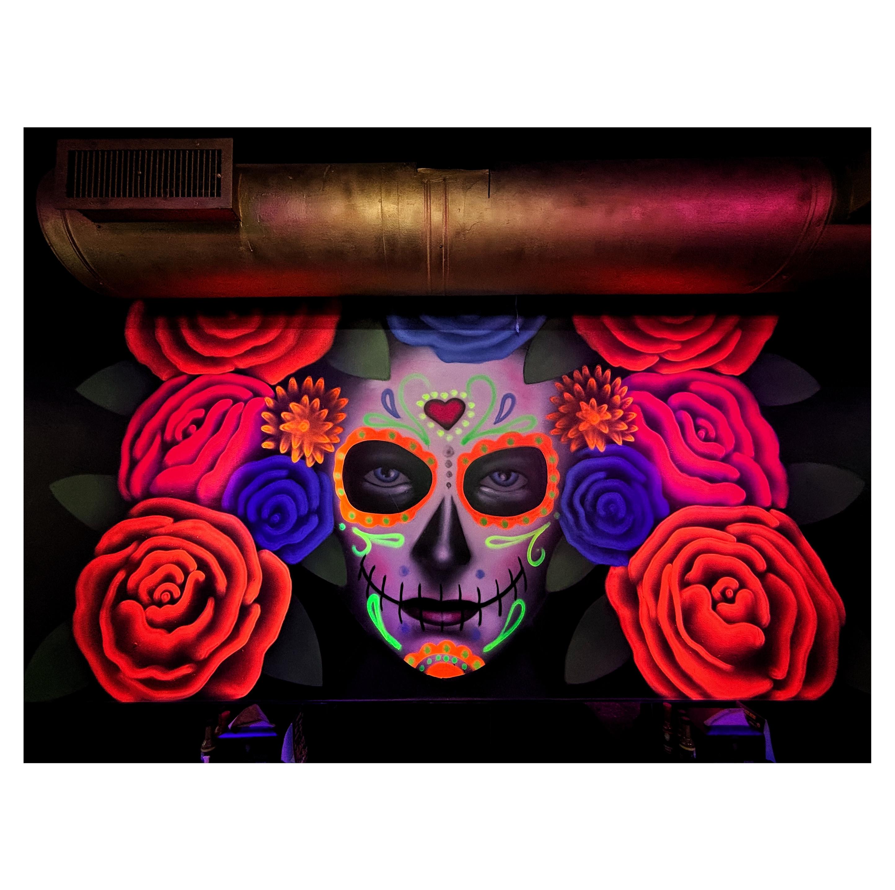 Photo of a mural done in vibrant glowing paint in a darkened restaurant interior. The mural depicts a feminine face in colorful makeup evoking a Mexican Day of the Dead calavera (skull), surrounded by giant roses in vivid red and purple. Mounted on the wall above the mural is a fat horizontal tube with a rectangular vent for air conditioning.