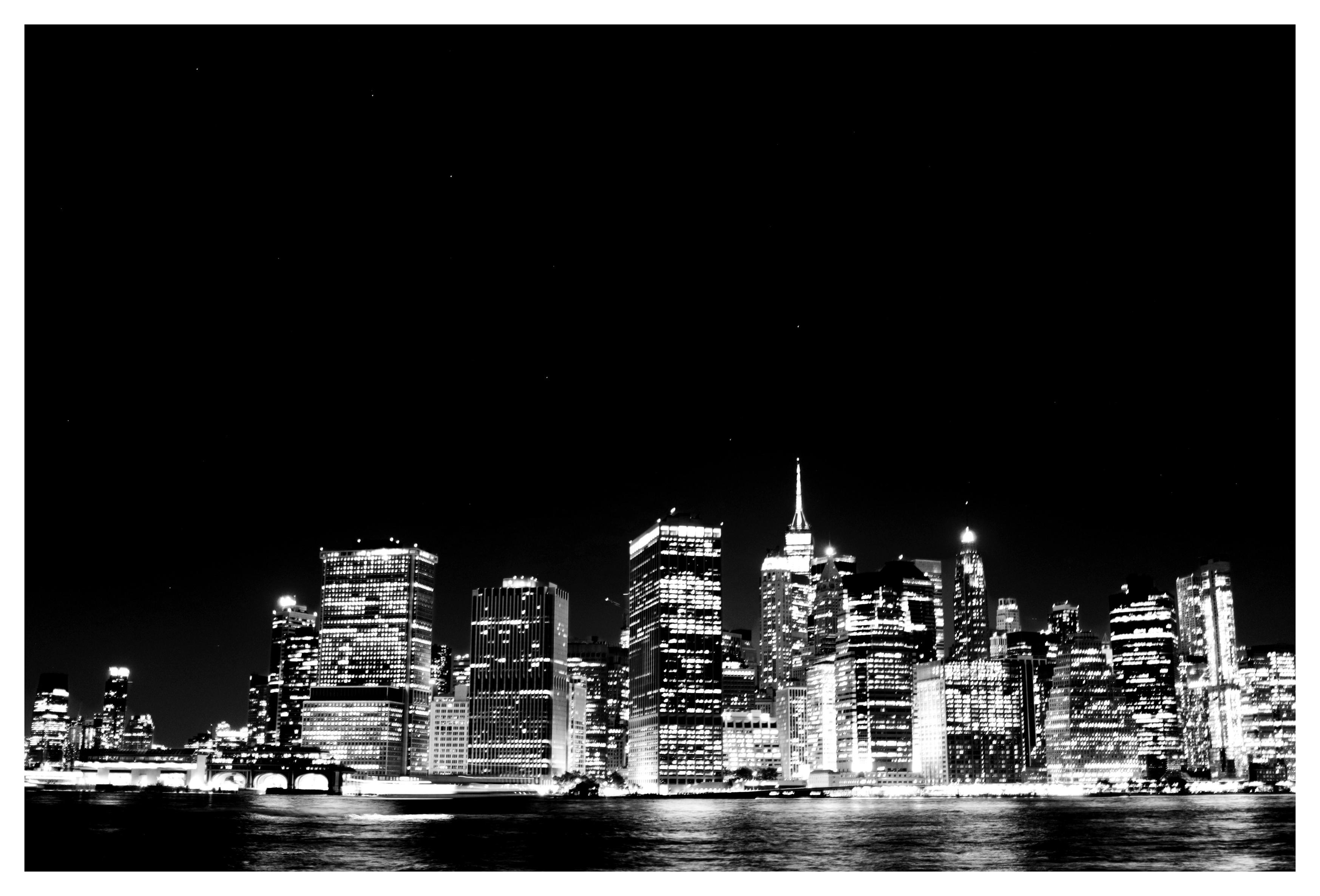 High-contrast black and white photo of the Manhattan skyline at night, its millions of windows illuminated, with the East River in front of it reflecting the city’s glow. Above is a completely black sky with a few faint stars: the constellation Ursa Major (the Great Bear or Big Dipper).