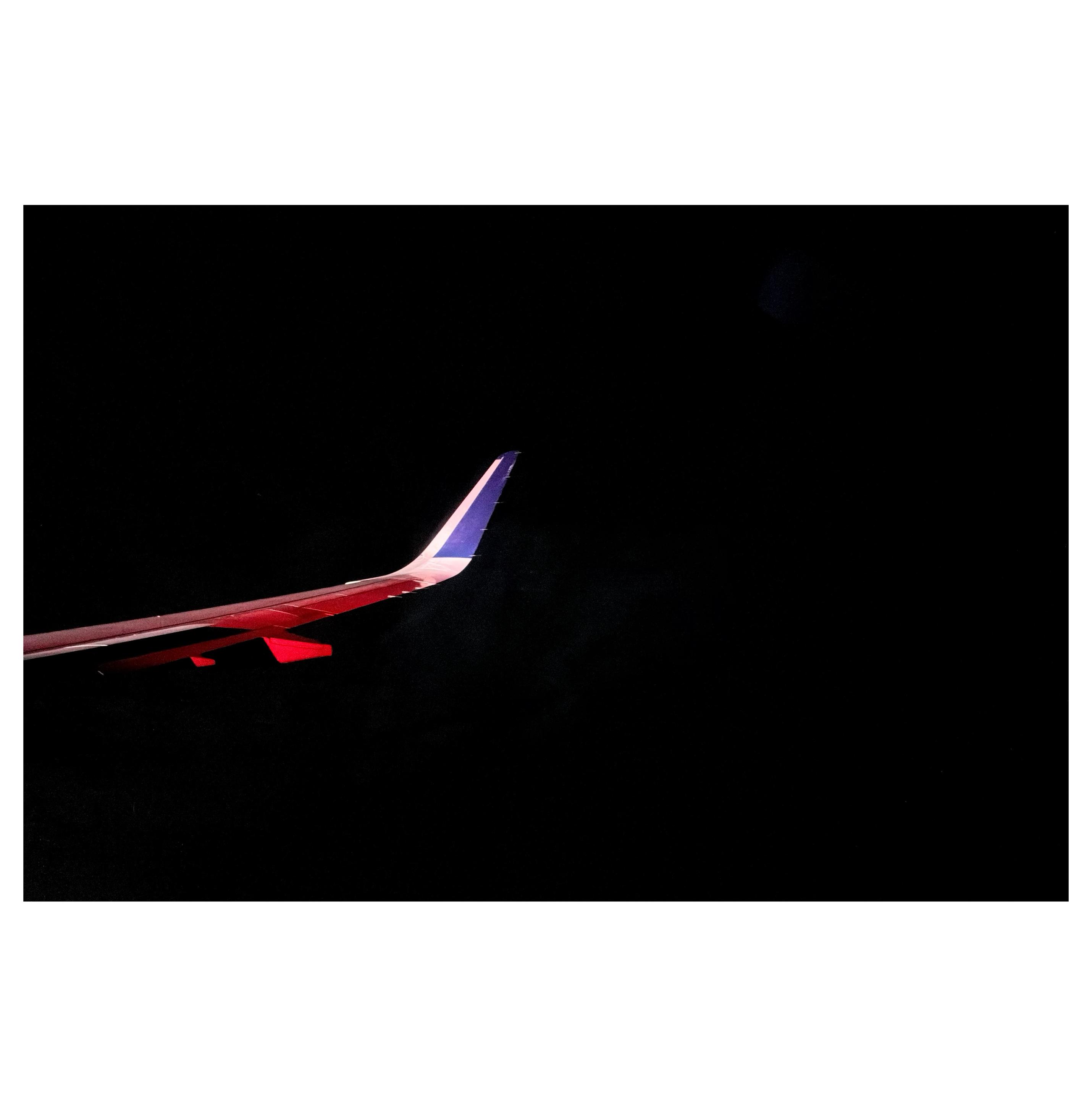 A view, outside an airplane window, of the plane’s right wing against an almost absolutely black night sky with just the faint ghosts of clouds. The wingtip is bent upwards with a blue stripe on a white field. A large section of the wing glows red from a blinking signal light.