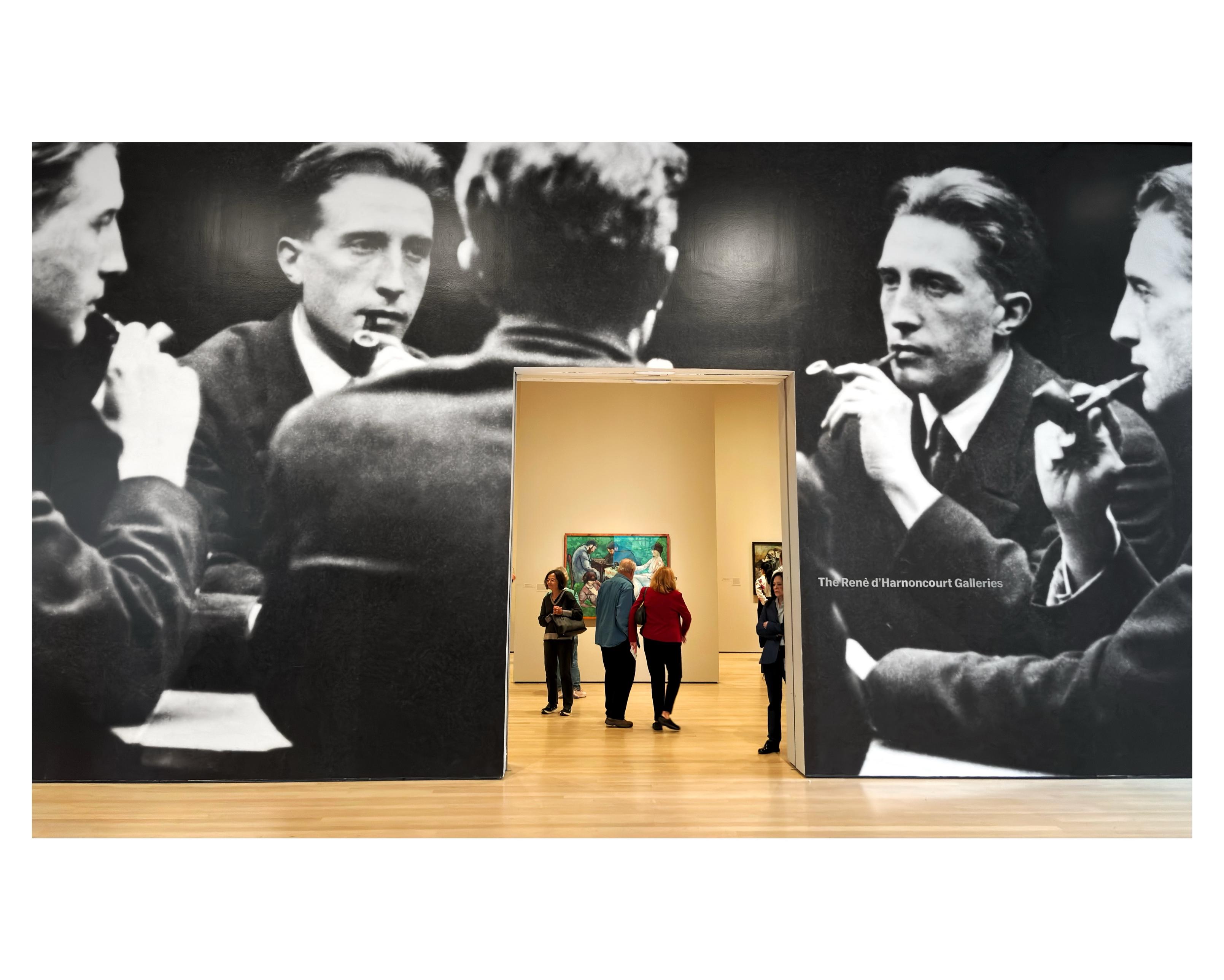 Photo taken at a Marcel Duchamp retrospective at the Museum of Modern Art in NYC. One wall is entirely occupied by a blown-up black and white photograph depicting Duchamp in a suit holding a pipe to his mouth and reflected in several mirrors, which gives the impression of identical businessmen seated around a table. The image dwarfs a doorway in the wall through which we can see museum visitors wandering about in another gallery.