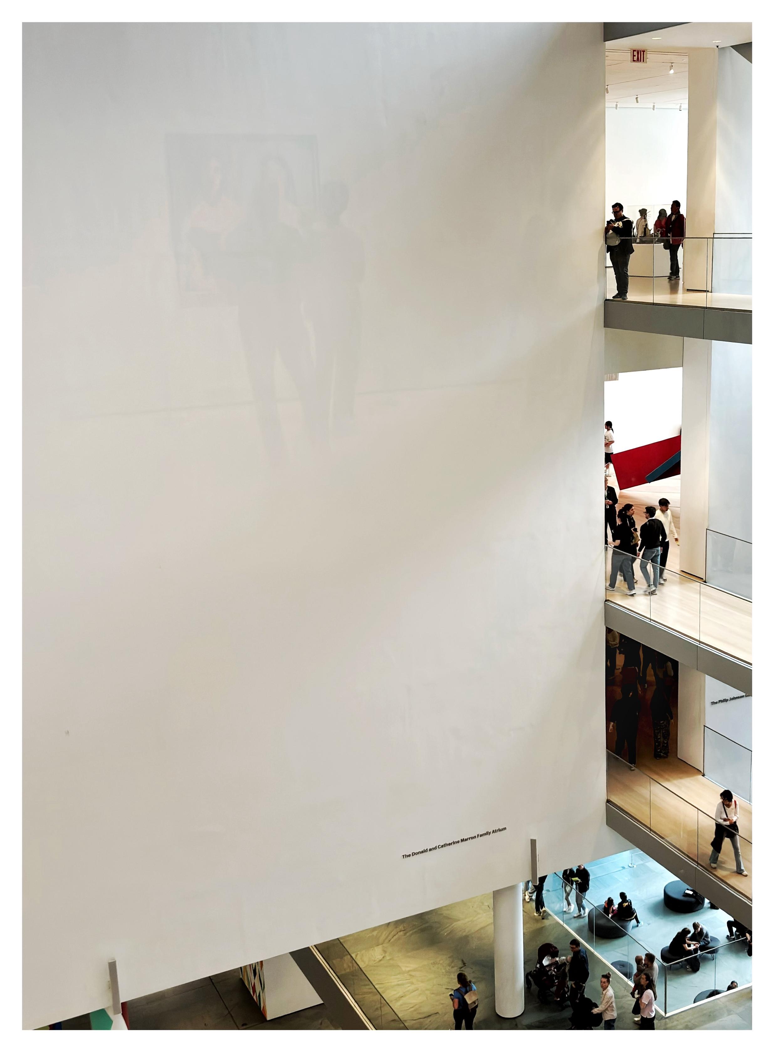 Photo taken from an upper balcony of the Museum of Modern Art in NYC, looking down at the main lobby. Occupying most of the image is a large blank white wall, supported by columns on the ground floor and spanning several levels as it rises up out of frame. A line of text near the bottom of the wall reads “The Donald and Catherine Marron Family Atrium.” On the right side of the photo we see people walking along three upper balconies perpendicular to the wall. At the bottom we see more visitors wandering around the ground floor, which is lit in warm light; through the glass panels of a railing we also glimpse a lower-level floor strewn with round cushioned seating, lit in colder blue light. The photo was taken through a glass barrier, and so on the white wall we can see a faint reflection of a couple perusing a painting behind camera.