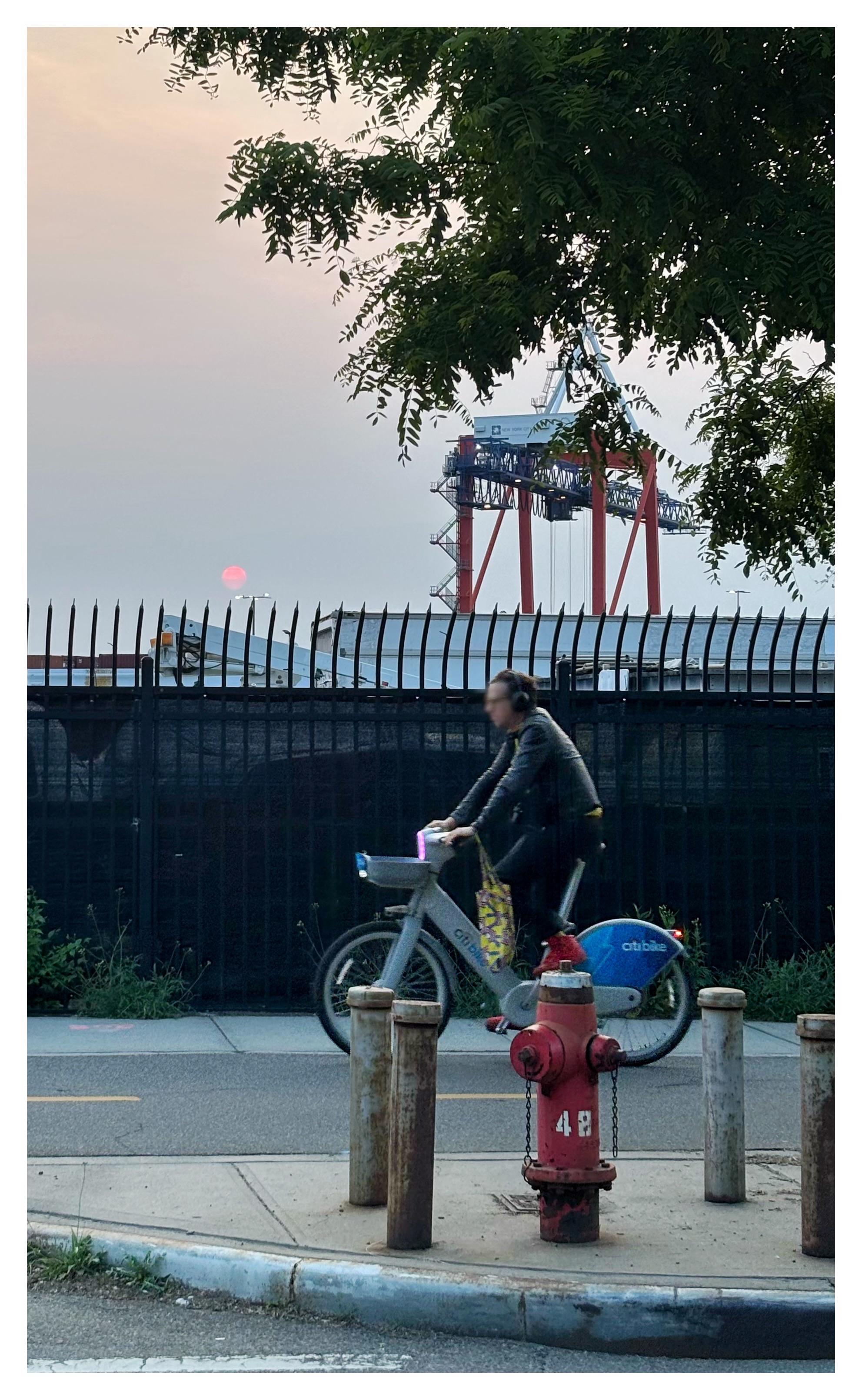 Sunset on a street near the Brooklyn waterfront. In the foreground is a street divider with a skinny red fire hydrant surrounded by four metal posts. The number “48” is spray-painted in white on the hydrant. On the far side of the street, a cyclist whizzes by on a blue-and-gray CitiBike. Behind him is a spiked fence obscuring some shipyard containers. A red-and-white shipping crane rises in the distance, partly obscured by leafy tree branches near the camera. The sky is a dull orange, with a small red sun dipping close to the fence — the colors resulting from smoke from the Canadian wildfires that day.