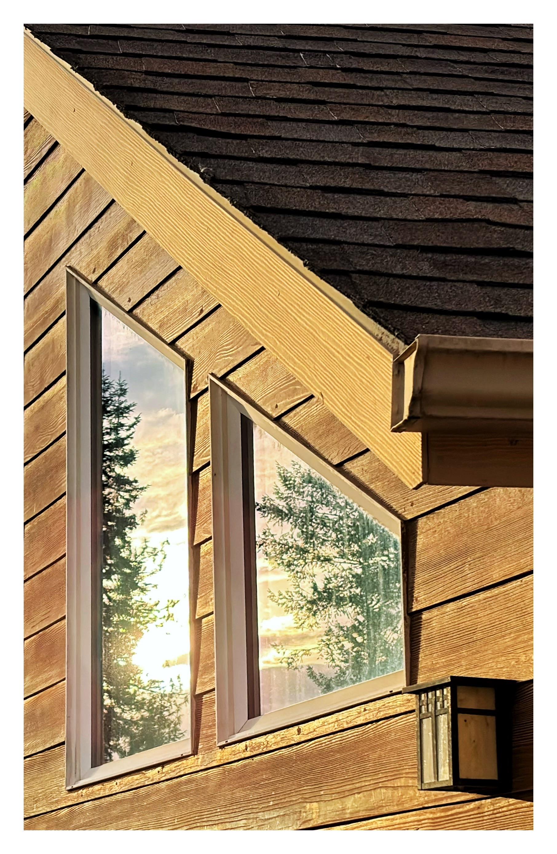 Closeup on two trapezoidal windows set in the clapboard siding of a house, their upper edges parallel to the slant of the shingled roof. A small rectangular lantern is mounted to the wall to the lower right of the windows. Fading daylight bathes the scene in a warm glow. The windowpanes reflect the sun setting between pine trees, brilliant in a golden sky.
